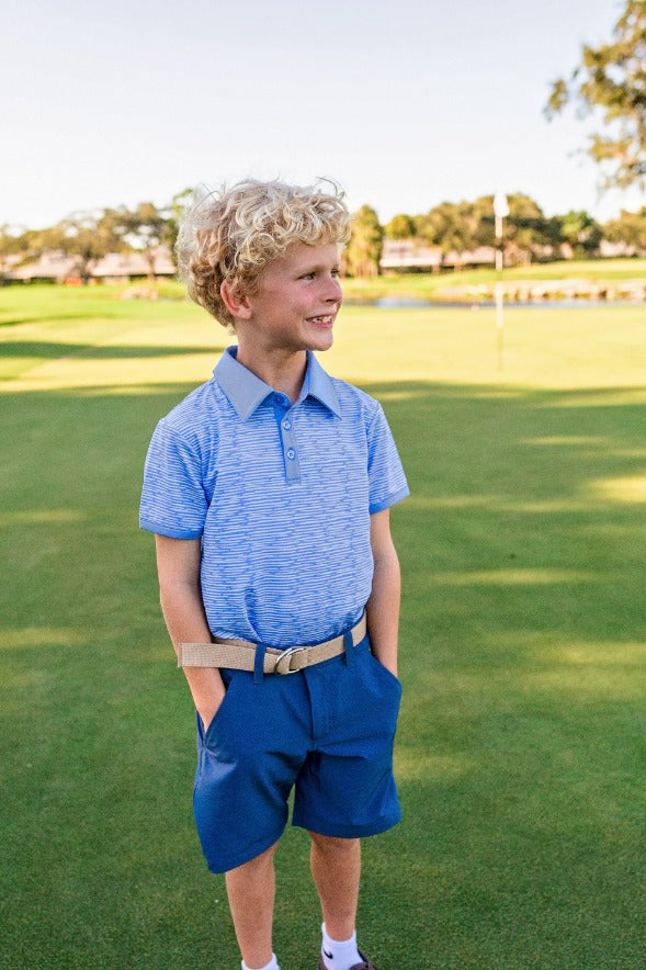 A young boy stands on a golf course wearing navy blue chino shorts and a light blue and white striped polo shirt