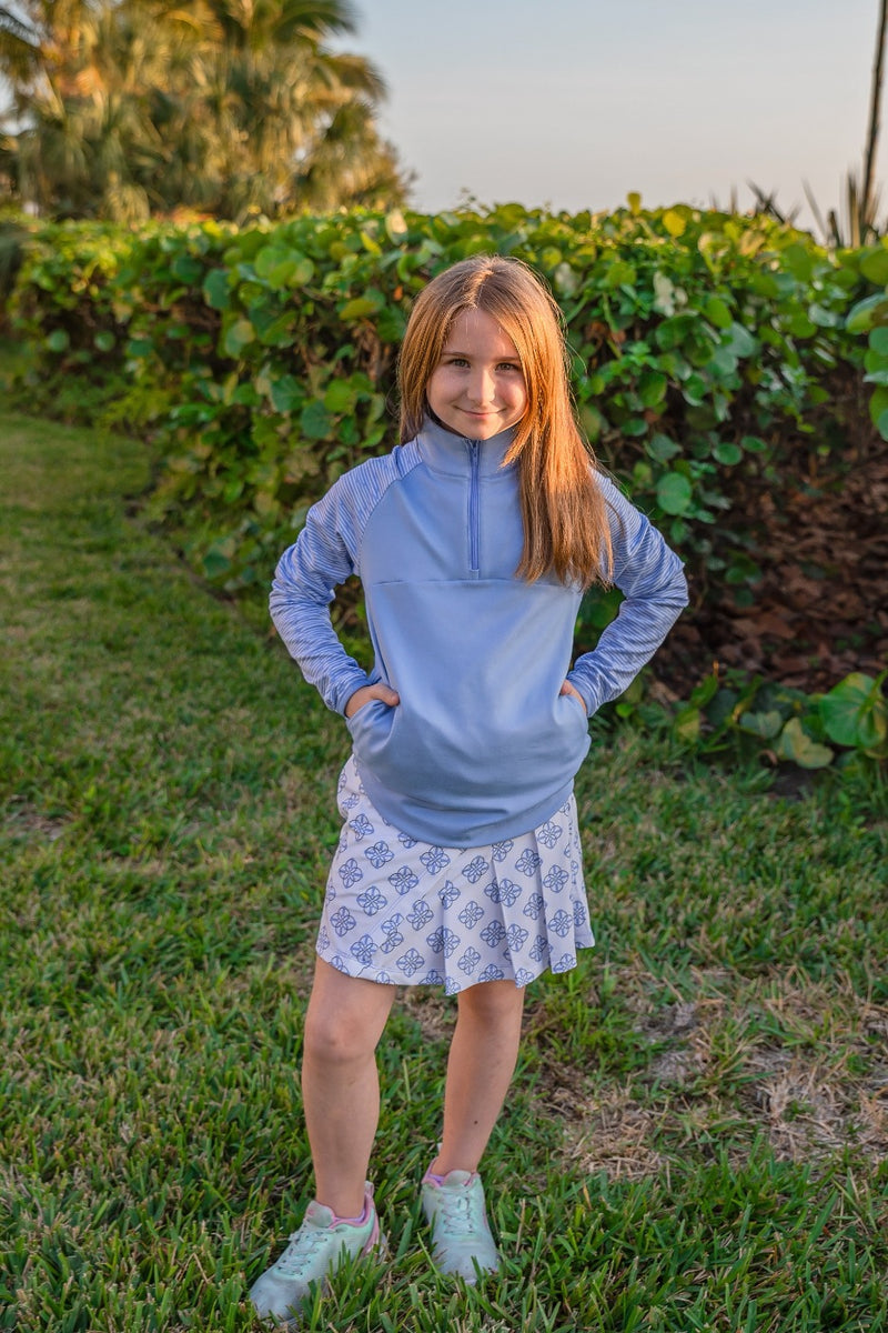 Young girl stands with her hands on her hips outside, wearing a blue quarter zip sweatshirt and a printed blue and white golf skort.