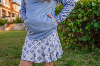 Young girl stands with her hands in her sweatshirt pocket outside, wearing a blue quarter zip sweatshirt and a printed blue and white golf skort.