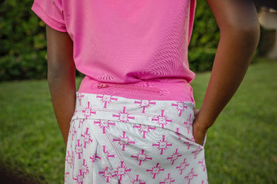 A young girl stands on a golf course modeling a sleeveless white polo shirt and a white golf skort with hot pink geometric prints and a hot pink waistband.