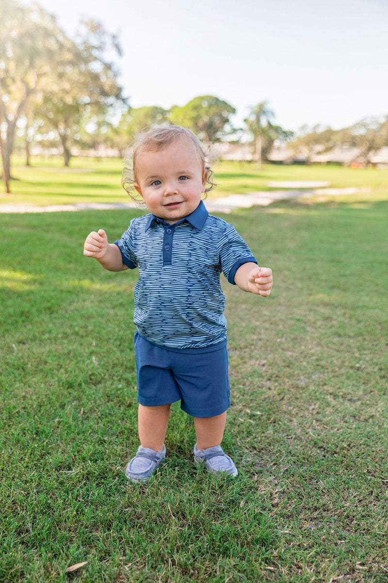 A young boy stands on a golf course wearing a navy blue and green polo shirt with a navy blue button up color. He is also wearing navy blue boys tennis shorts.